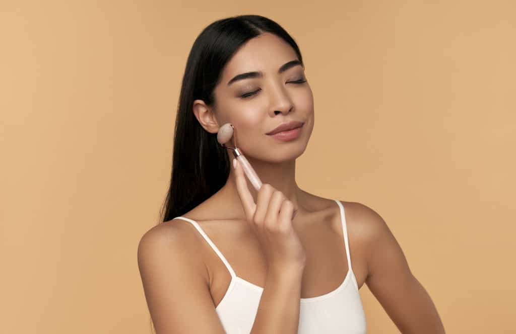 A young Asian woman with clean radiant skin makes a facial massage with a quartz roller on a beige background. Spa care, facial skin care, beauty cosmetology.